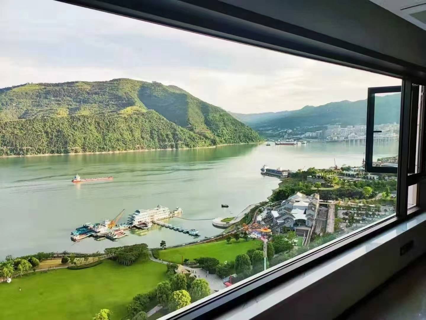 这是一张展示高层江景落地窗的实拍图。透过宽大的深色边框窗户，可以看到窗外壮阔的江面、连绵的青山以及岸边的绿地公园和码头船只。高透光的玻璃带来了极佳的采光和无遮挡