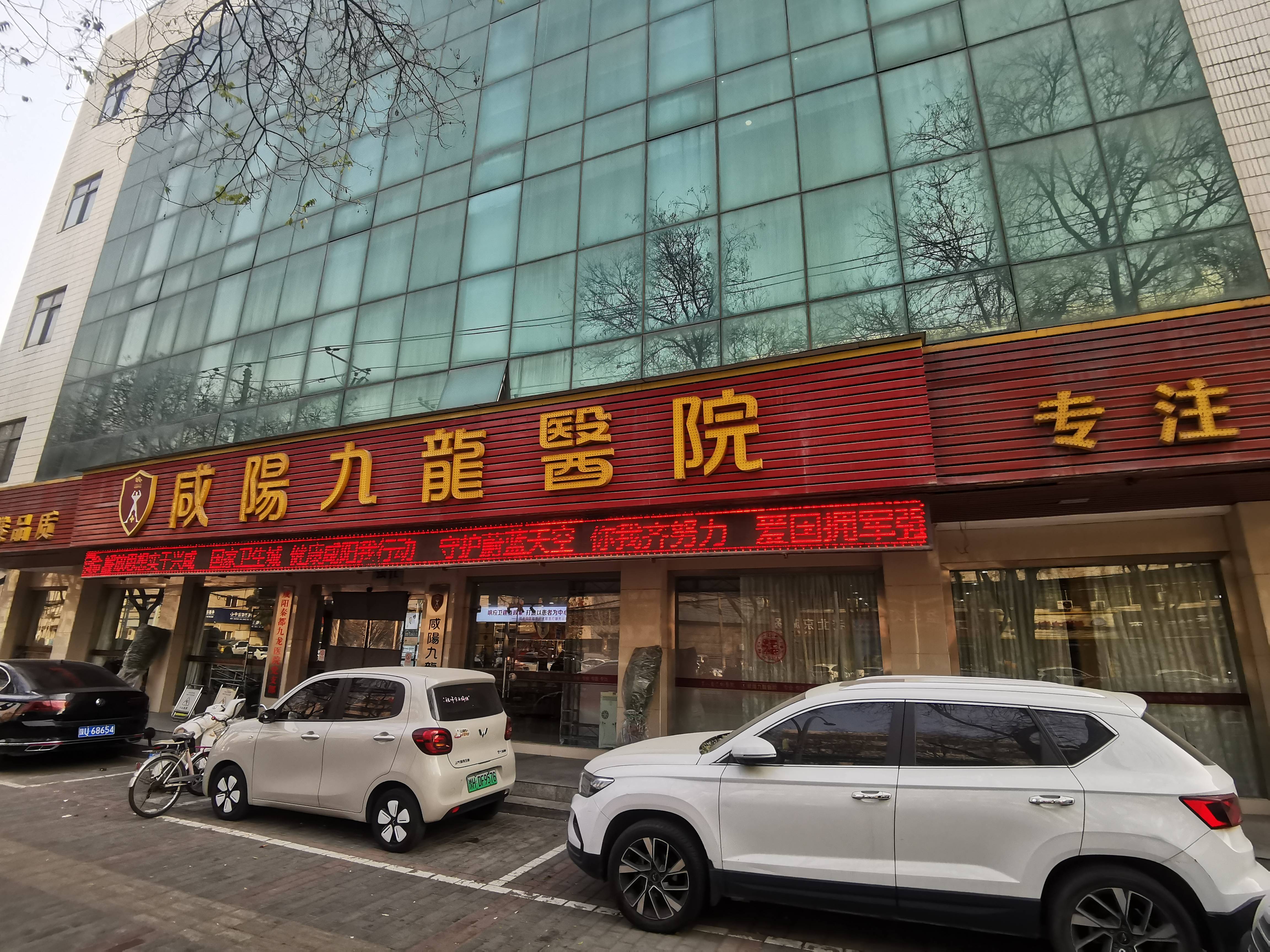 咸阳秦都九龙医院临街外观，电子屏显示公益宣传标语，门前停有多车