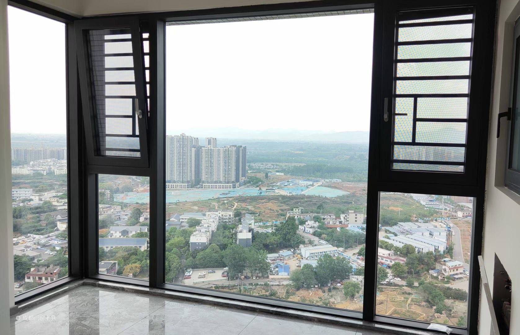 这张图片展示了高层住宅室内安装的一套黑色断桥铝全景落地窗。窗体采用深灰色铝合金型材，中间为大面积固定玻璃，两侧配有带金刚网纱窗的开启扇，兼顾通风与防蚊安全。窗外